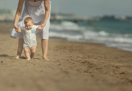 A little boy learns how to walk on the sand with support of his mother