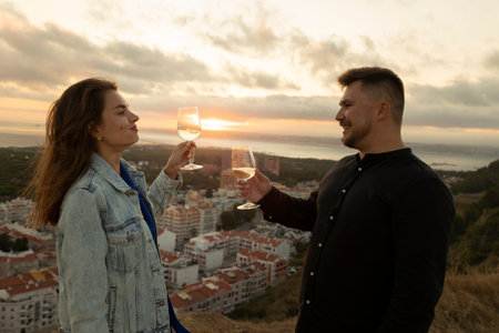 Young couple clinking glasses with white wine, enjoying scenic sunset over cityscapeの写真素材