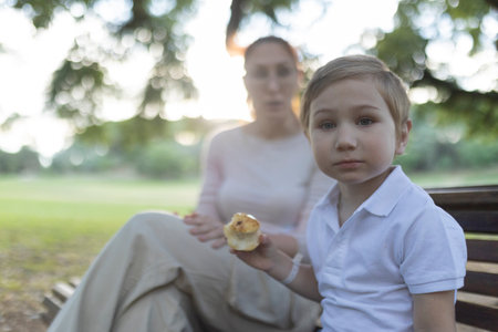 Young boy eating apple sitting on bench with mother in parkの写真素材