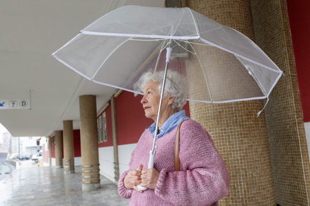 Elderly woman holding transparent umbrella walking in rainy cityの写真素材