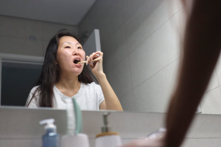 Young woman brushing teeth with wooden toothbrush in bathroom mirror reflectionの写真素材