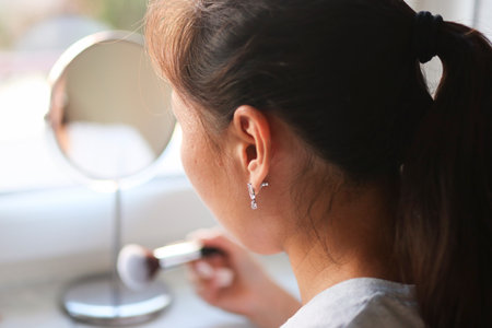 Woman applying makeup with brush near mirror in natural lightの写真素材