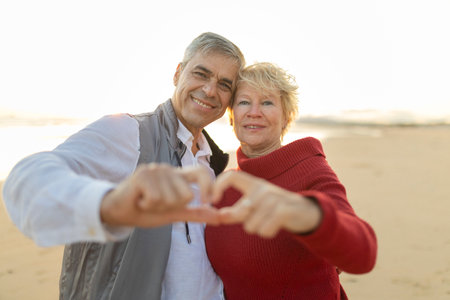 Happy senior couple making heart shape with hands on beach at sunset