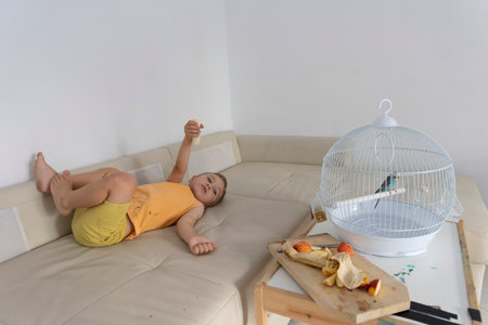 Child lying on sofa sharing banana with pet budgie in cageの写真素材