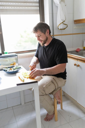Man sitting on stool cutting zucchini in home kitchenの写真素材