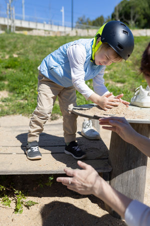 Child playing with sand at playground wearing helmet and watched by therapist