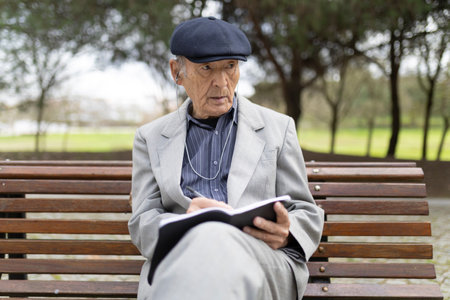 Senior man writing notes while listening music on bench in parkの写真素材