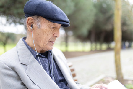 Elderly man listening music with earphones and reading book in park
