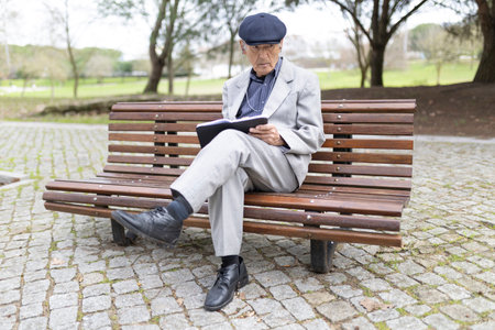 Senior professor reading book while sitting on bench in parkの写真素材