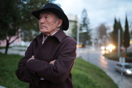 Pensive senior man wearing hat standing with arms crossed in parkの写真素材