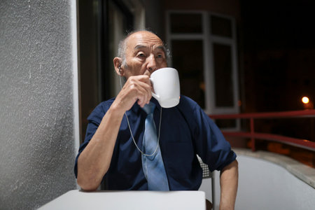 Senior businessman drinking coffee on balcony at night listening to musicの写真素材
