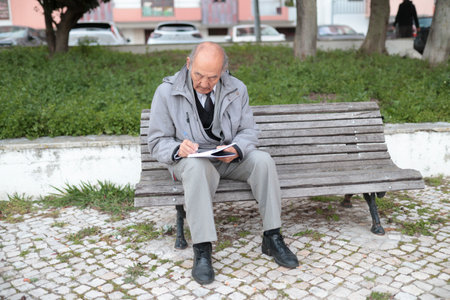 Senior professor writing notes sitting on bench in university campus parkの写真素材