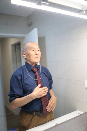 Senior businessman adjusting tie and using hearing aid in bathroom mirrorの写真素材