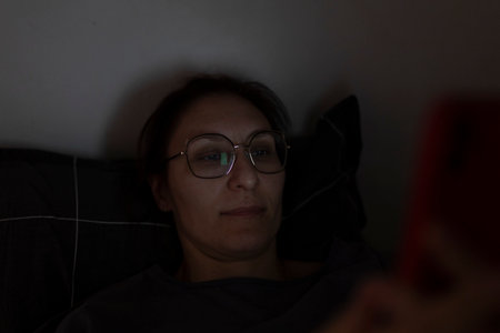 Woman wearing glasses using smartphone in bed at nightの写真素材