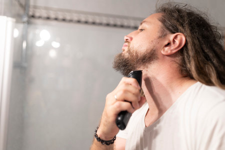 Young man trimming his beard with electric razor in bathroom mirrorの写真素材