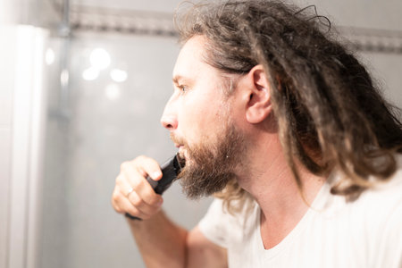 Man trimming his beard with electric razor in bathroom mirrorの写真素材