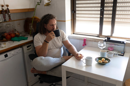 Man drinking coffee and eating biscuits in messy kitchenの写真素材