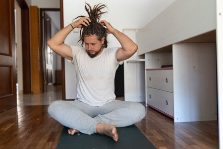 Young man with dreadlocks fixing his hair while sitting on yoga mat in bedroomの写真素材