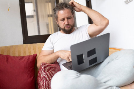 Confused freelancer scratching head while working on laptop at homeの写真素材