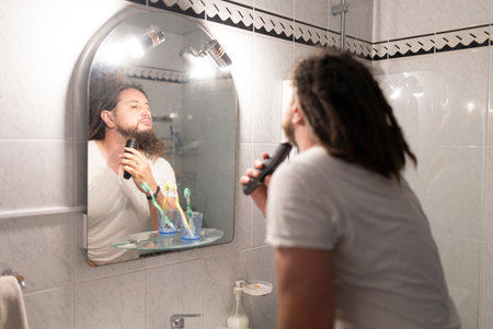 Young man shaving beard with electric razor in bathroom mirrorの写真素材