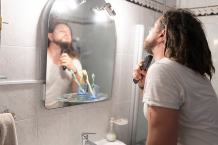 Young man shaving beard with electric razor in bathroom mirrorの写真素材