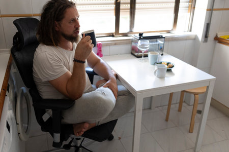 Man drinking coffee in home kitchen sitting on office chairの写真素材