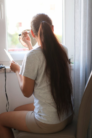 Woman applying makeup while reading instructions in front of windowの写真素材