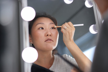 Asian woman applying makeup with brush in front of mirror with lightsの写真素材