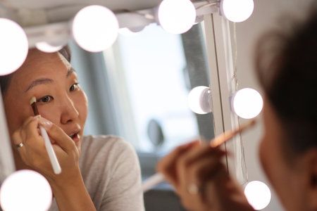 Asian woman applying makeup in front of mirror with lightsの写真素材