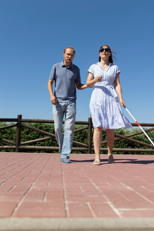 Man guiding blind woman with white cane walking in parkの写真素材
