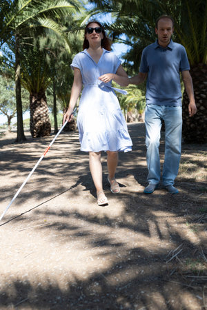 Blind woman walking with white cane and friend in parkの写真素材