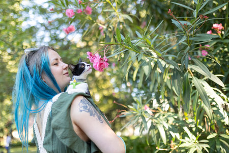 Woman with blue hair holding cat smelling pink flowers in a gardenの写真素材