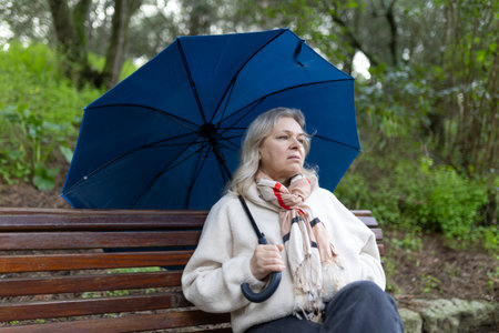 Pensive senior woman holding umbrella sitting on park bench on rainy dayの写真素材