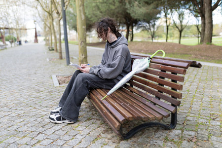 Young man writing in notebook sitting on park bench with umbrella