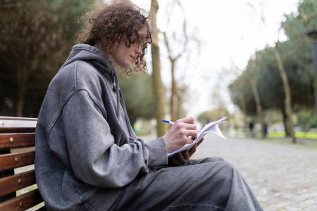 Young man writing notes in notebook while sitting on bench in park