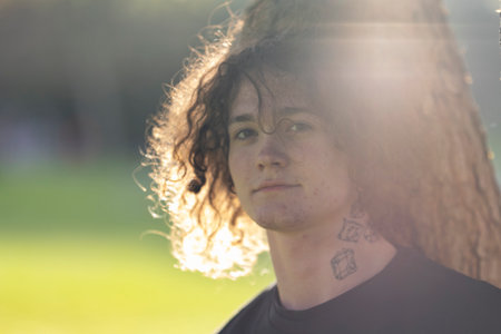 Young man with curly hair and tattoos posing outdoors in sunlight