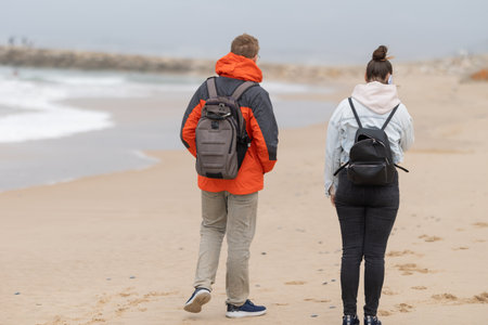 Two people walking beach observing ocean wearing backpacks