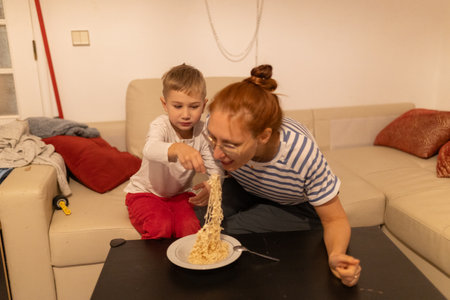 Boy feeding instant noodles to mother at homeの写真素材