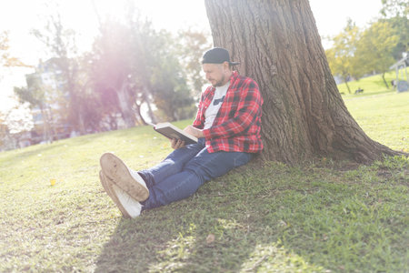 Man relaxing in park, reading book under treeの写真素材