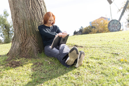Woman relaxing outdoors reading book under treeの写真素材