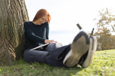 Woman relaxing park reading book leaning against treeの写真素材
