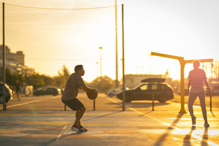 Young adults playing basketball on outdoor court at sunset