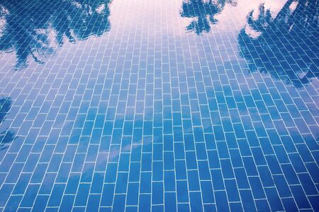 Blue tiled floor of a pool under clear water, in which sky and palm trees are reflected