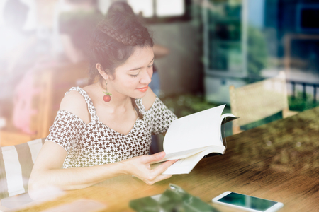 Young woman enjoying reading book sitting near window relaxing in her living with window reflection in the coffee shopの写真素材