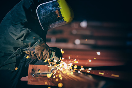 Worker with the face guard to electric wheel grinding on steel structure in factory. add grain filter dark tone heavy work concept.の写真素材