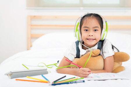 Portrait asian little girl listen to music from tablet with green headphone while writing with colorful pencils on bed. Education for kids and strengthens the imagination of children conceptの写真素材