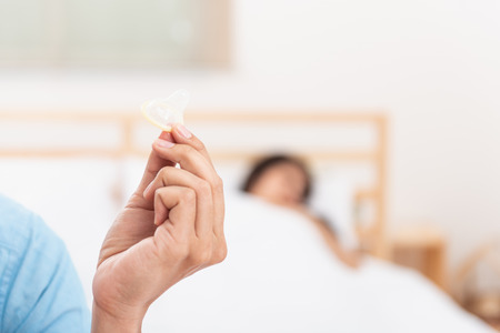 Close Up of a hand of the young man holding opening condom with a woman on the bed is background. Focus to a condom.  Best condom for protectionの写真素材
