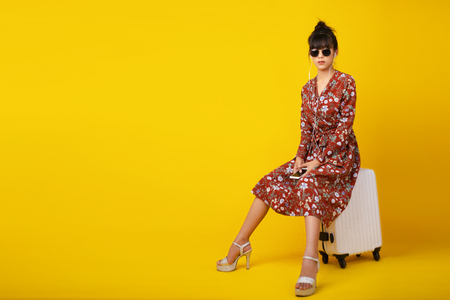 Asian young woman in vintage dress and wearing sunglasses sitting on white suitcase while her earphone on yellow background. Lifestyle, Fashion and travel concept.の写真素材