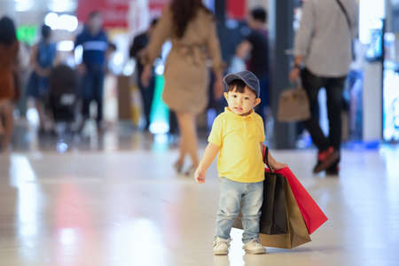 Kid shopping. Asian little boy in yellow shirt and jeans enjoyment of shop. Walk in the shopping mall and holding heavy shopping paper bags.の写真素材