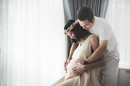 Happiness pregnant woman sitting relaxed with husband are hugging and smiling while standing near the window at home Moments for relationship of pregnant couple.Waiting for baby.の写真素材